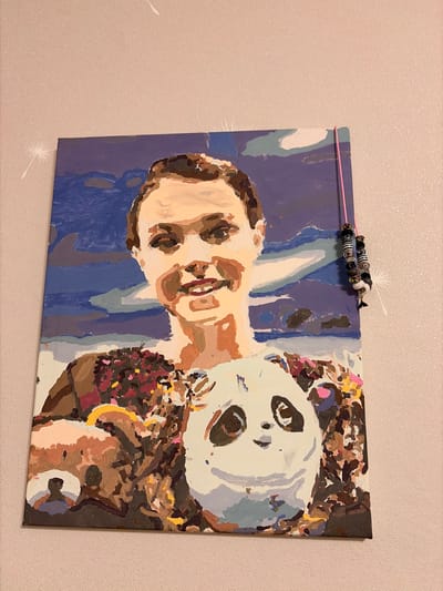 A painting of a person holding a panda is displayed on a wall. The painting is in a cubist style with bold blocks of color. The person in the painting has short brown hair and is smiling. They are wearing a brown shirt. The panda is white with black markings. The background of the painting is a blue sky with white clouds. The painting is hanging on a light pink textured wall. A pink string with beads is attached to the top right corner of the painting and hangs down the side. There are two white dandelion-like designs on the wall to the left of the painting. The painting appears to be in a domestic setting. There is no identifiable information about Leninogorsk, Russia in the image.