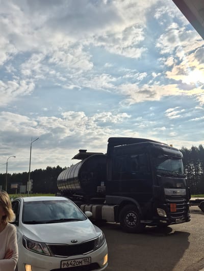 In the city of Mozhary, Russia, a black DAF tanker truck and a white Kia Rio are parked on an asphalt surface. The tanker truck is positioned to the right of the Kia, with its trailer extending behind it. A woman stands to the left of the Kia, with her arms crossed. The sky above is filled with a mix of clouds and sunlight, suggesting it is daytime. In the background, a line of trees forms a dark silhouette against the horizon. The scene appears to be a roadside or parking area.