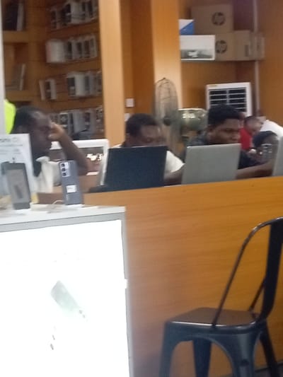 Three individuals are seated behind a counter, each using a laptop. Displayed are various electronics and packaging for HP products. A black metal chair is visible in the foreground. A fan is present in the background. The walls are a warm orange color.