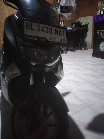 A Yamaha motorcycle with license plate BL 3430 NAI, expiring 07-29, is parked indoors. The background contains indistinct furniture and objects. The scene is presumably located in Kruengmanyang, Indonesia.
