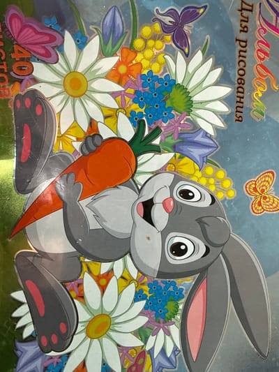 This image displays a brightly colored, digitally rendered illustration, likely from a children's album or activity book, observed in the city of Apnagyugh, Armenia. The central subject is a cartoon grey rabbit, depicted lying on its back, holding an orange carrot with green foliage in its paws above its head. The rabbit features large black eyes with white pupils, a pink nose, and an open-mouthed expression revealing a small pink tongue. Its long grey ears contain pink interiors, and its paws have black pads with red accents. The background consists of a dense, multi-colored floral arrangement, including large white daisies with yellow centers, blue forget-me-nots, yellow clusters resembling mimosa, orange flowers, and purple bell-shaped flowers. Two butterflies are also present: a larger purple one in the upper right quadrant and a smaller yellow-orange one in the lower right. Cyrillic text is visible; "Альбом для рисования" (Album for drawing) is vertically oriented on the right side, and "40 листов" (40 sheets) is partially visible on the left side. The image itself is a photograph of a physical object, exhibiting surface reflections and minor indentations on the rabbit's face.