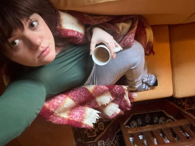 A woman in a green long-sleeved shirt and gray sweatpants lies on a tan couch, covered with a red and cream plaid blanket. She holds a white mug with coffee in her right hand. Her brown hair is parted to the side, and she gazes upward with a slight smile. Above her, a dark rug and a wooden table with ornate legs are visible. The overall ambiance is cozy and relaxed.