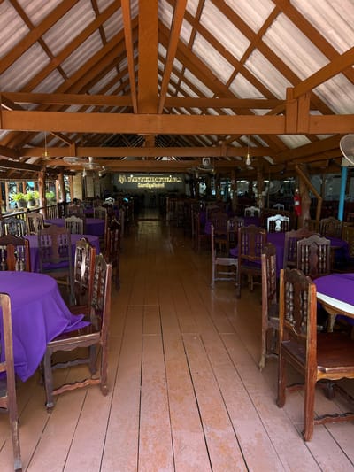 This is a ground-level, interior shot of a restaurant located in Tambon Ko Panyi, Thailand. The restaurant has a wooden structure with a high, exposed wooden beam ceiling and a corrugated roof. The dining area is furnished with numerous wooden chairs and round tables covered with vibrant purple tablecloths. The floor is made of wooden planks. In the background, a bar area is visible with stools and patrons. Various ceiling fans and lighting fixtures are also present. The overall ambiance suggests a casual, open-air dining establishment.