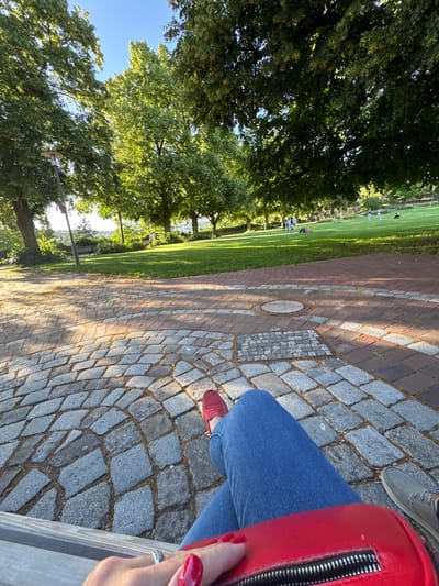 This is a first-person perspective shot of a person sitting on a bench in Esslingen am Neckar, Germany. The foreground is dominated by the cobblestone pavement and the person's legs clad in blue jeans and red loafers, with a red handbag resting on their lap. The middle ground features a pathway with circular brickwork and a manhole cover. In the background, a lush green lawn is dotted with several people enjoying the outdoors. Towering trees with dense foliage cast dappled shadows across the scene, and a clear blue sky suggests a pleasant day. The overall setting appears to be a park or a scenic viewpoint overlooking the city.