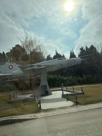 The image shows a gray, retired MiG-21 fighter jet, a common aircraft model for the Soviet military. The jet has a red "277" painted on its side and a red circle with a white star in the center on its tail. It is mounted on a concrete stand in a grassy area off the side of a paved road, likely a small town road, in Sunny Beach, Bulgaria. The jet is angled upward towards the sky and a bright sun is shining in the partly cloudy sky. The jet is surrounded by trees, likely pines, and a willow tree.