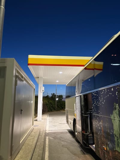 The image shows a Shell gas station at dusk in Kalotina, Bulgaria. A long bus with images of the Statue of Liberty and buildings of New York City is parked beside the gas station. The sky is a deep blue, and the gas station canopy is lit from underneath. A small white building is in the foreground. A number 8 is on the post of the gas station.