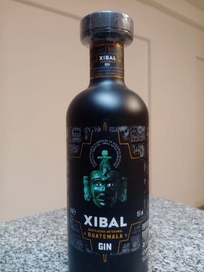 A single, sealed bottle of Xibal Guatemala Gin stands upright on a speckled grey and white countertop, likely granite. The bottle is matte black with a label featuring a prominent teal-colored stylized mask design, identifiable as Mesoamerican, above the text "XIBAL GUATEMALA GIN" and "DESTILACIÓN ARTESANAL." The black bottle cap is secured with a clear plastic shrink wrap, indicating it is unopened, and shows visible perforations. The background consists of a plain, light beige wall with subtle vertical panel lines. No individuals are present. The scene is located in Lomas de Zamora, Argentina.
