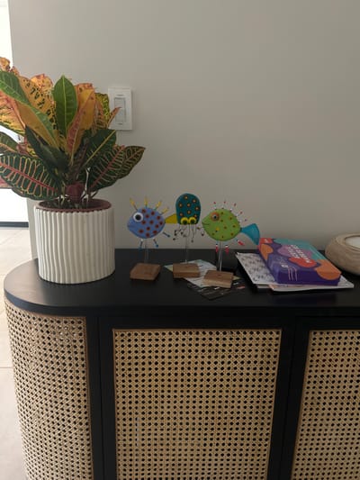 The image shows the interior of a home in Playa Herradura, Costa Rica, at the coordinates 9.658 latitude and -84.673 longitude. There is a black cabinet with woven rattan panels on the front. The cabinet has a black top surface, where three colorful glass fish figurines are arranged, one next to the other. The figurines are on wooden stands. There are also some papers and a purple box with the words "Acrylic Markers" on it. A large potted plant with colorful, patterned leaves is located to the left of the cabinet. The plant is in a white ceramic pot with vertical grooves. A white light switch is located on the wall behind the plant. The background is a white wall. The image is taken from a slightly elevated angle, looking down at the cabinet. The lighting in the room is bright and evenly distributed.