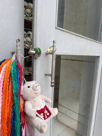 A white door with a glass pane stands ajar, revealing shelves filled with human skulls in what appears to be a memorial or museum setting. In the foreground, a string of colorful origami cranes hangs from a metal bar on the door, alongside a plush pig toy wearing a red and white garment. A small stuffed koala, wearing a green vest with "I AUSTRALIA" written on it, is attached to the door handle by a string. The scene suggests a somber yet decorative display at a location in Khan Dangkao, Cambodia, likely related to historical events.