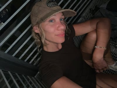 The image shows a person lying down, wearing a brown t-shirt and a brown and beige patterned baseball cap. They have blonde hair and blue eyes and are wearing a bracelet on their left wrist. The person appears to be outdoors in Ohlstadt, Germany, as indicated by the presence of a numbered sign (partially visible as "578") on a metal railing, typical of park or recreational areas. The background consists of metal railings and dark foliage. The lighting suggests it is either evening or nighttime.