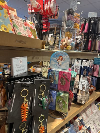 The image shows a retail display of small gift items, primarily keychains and socks, arranged on shelves and hanging racks in a store. The items are predominantly adorned with German text and playful graphics, suggesting a focus on the German gift market. Keychains feature various charms like angels, animals, and fish skeletons, some presented with individual cards that include descriptive text such as "Schutzengel" (guardian angel) and "Glück auf deinen Wegen" (luck on your path). There are also displays of socks packaged in blister packs, with slogans like "Zauber-Socken" (magic socks). In the background, more shelves are visible with a variety of merchandise, including small stuffed toys. The overall impression is of a well-stocked gift shop. The presence of German language and the specific types of novelty items are consistent with a shop in Germany. Given the context of the available metadata, this scene is likely from a store in the city of Herford, Germany.