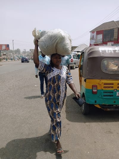 The image depicts a daytime street scene in Kadoma, Zimbabwe. In the foreground, an adult female of African descent is walking towards the viewer, positioned slightly to the left of the frame's center. She is wearing a long, full-length dress with a blue, brown, and white patterned design, and open-toed sandals. She is carrying a large, bulky, off-white sack-like package balanced on her head, supported by her raised right hand. Her left hand holds a dark, rectangular object resembling a wallet or clutch. Her facial expression is neutral, and she is looking directly forward.

Behind the woman, two other adult males are visible walking on the asphalt road; one wears a light blue hooded top and dark trousers, and the other a light-colored shirt. To the right of the main subject, a yellow and green three-wheeled motorized rickshaw (auto-rickshaw) is present on the roadside. Behind the rickshaw, a building with a brown-tiled roof is visible, featuring a prominent red sign with white text, including "YANGA O.X.". Overhead wires run across the upper right portion of the frame.

To the far left of the road, a dark blue car is visible in the distance, followed by other distant vehicles and commercial buildings. A red billboard is partially visible on the far left. The sky is bright and appears somewhat hazy. Direct sunlight casts shadows on the asphalt road, with the woman's shadow extending behind her. The road surface shows signs of wear.
