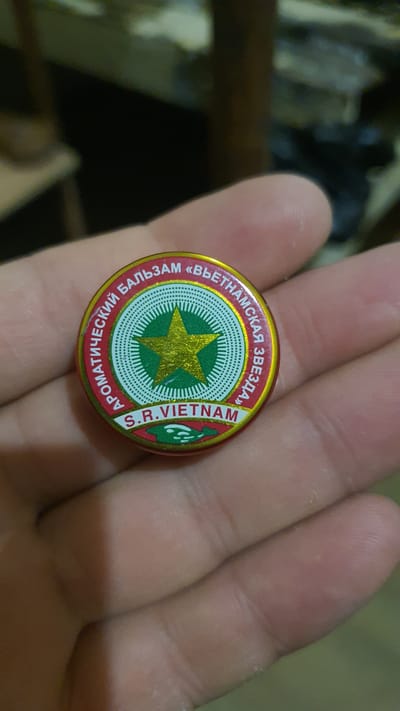 A person's hand is holding a small, round, metallic tin with a red and gold label. The label features a green circular design with a gold star in the center, surrounded by radiating lines. Text in a circular pattern reads "АРОМАТИЧЕСКИЙ БАЛЬЗАМ «ВЬЕТНАМСКАЯ ЗВЕЗДА»" and "S.R.VIETNAM". Below the circular design, there is a green band with a white, stylized depiction of a fish. The background is blurred and appears to be an indoor environment, possibly a room, with muted lighting. The image does not provide any information about the location in the city of Karankul, Uzbekistan.