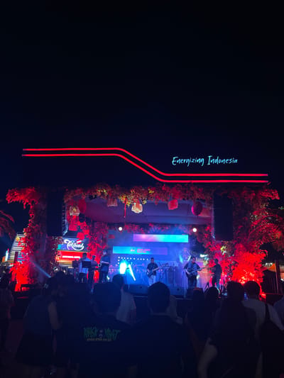 The image depicts an outdoor musical event in Salembaran Jati, Indonesia. A stage adorned with red foliage is illuminated by stage lighting. A band performs on stage in front of an audience. The backdrop includes "Energizing Indonesia" written vertically on the left of the image and several light elements. Audience members are primarily seen from behind, suggesting they are focused on the performance. A lit sign that says "teh Botol" is prominently displayed at the bottom.