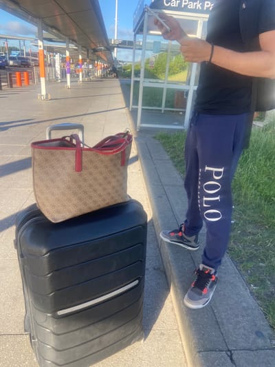This image depicts a person standing next to luggage at a bus or coach station in Uttlesford, United Kingdom. The person, wearing dark blue sweatpants with "POLO" written in white lettering and grey and red sneakers, is holding a smartphone. Beside them is a large black suitcase and a patterned tote bag with red trim. The background shows a covered waiting area with benches, supporting pillars, and signage, as well as cars and other people in the distance. The ground is paved, and there is a grassy area to the right. The lighting suggests it is daytime, likely a sunny day.