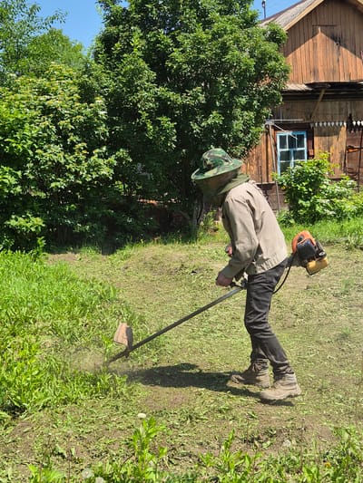 A person wearing a camouflage hat with a protective net, a light-colored jacket, dark pants, and sturdy boots operates a string trimmer in a grassy yard. The string trimmer is powered by a small engine attached to its body and has a long shaft. The person is actively trimming the grass, which is creating a visible cloud of dust and cut vegetation. In the background, a wooden house with a blue-framed window and visible wooden siding stands under a clear blue sky. Lush green trees and bushes surround the house and yard, suggesting a rural or suburban setting in Novosisoevka, Russia. The ground is a mix of mown grass and uncut vegetation.