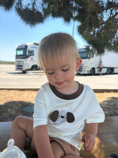 A young child with blond hair and fair skin is shown from the chest up, seated outdoors at a wooden table. The child is wearing a white t-shirt with a brown collar and a graphic of a koala on the front. They are also wearing brown shorts. The child is looking down and appears to be eating a piece of apple.
In the background, two white semi-trailer trucks are parked. One truck is closer and more in focus, with a person standing near the driver's side door. The other truck is further in the distance. Trees with green foliage are visible to the left, and the sky is blue with no visible clouds. The lighting suggests it is daytime, likely sunny. The setting appears to be a rest stop or parking area for trucks, possibly in Travel, France, given the context. The child's expression is one of gentle focus on the food.
