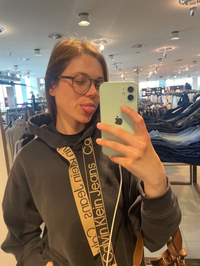 A woman wearing glasses and a dark Calvin Klein sweatshirt is taking a selfie with a light green iPhone. She is sticking her tongue out slightly. The background shows a clothing store with racks of jeans. The location may be in the city of Keszthely, Hungary.