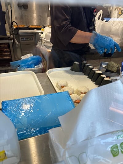 The image shows a person wearing blue gloves, preparing food, possibly in a commercial kitchen or restaurant in Chiyoda City, Japan. The person is wearing a black shirt and blue gloves, and is holding a piece of seaweed, likely for making rice balls. The person is standing behind a metal counter with several white plates containing rice balls wrapped in seaweed and some with pink filling. The plate contains multiple rice balls in a row, some covered in seaweed and others with pink filling, presumably sausage. Next to the plates is a blue plastic sheet, possibly for food preparation. The background shows a white plastic bag with a green logo, possibly a brand of food or packaging materials, and some blurry objects, possibly food items. The environment appears to be a kitchen or food preparation area. The person seems to be preparing the rice balls for sale or to be served to customers. The location of the scene is 35.681 latitude and 139.768 longitude, which places the scene in the Chiyoda City, Japan.