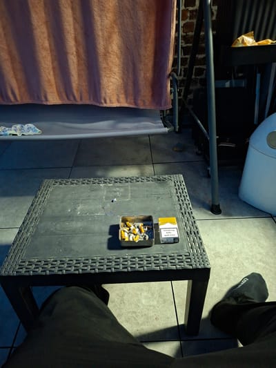 An outdoor scene, likely a patio, in Evere, Belgium, features a dark grey, woven-pattern plastic table in the foreground. On the table sits a metal ashtray filled with numerous cigarette butts and a pack of Marlboro cigarettes with a warning label. To the right of the table, partially visible, is a white trash receptacle. In the background, a pink towel hangs from a metal structure. The ground is tiled. The bottom of the image shows a person's legs and feet, one with a black sock.