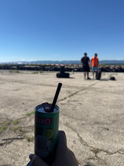 In Buhovtsi, Bulgaria, a person holds a can of "XIXO Mojito Lemonade" with a black straw. In the background, two men stand near a tire barrier, one wearing an orange life vest, suggesting an outdoor recreational area, possibly a go-kart track or similar venue. The ground is concrete with some grass patches. The sky is clear and blue, indicating fair weather.