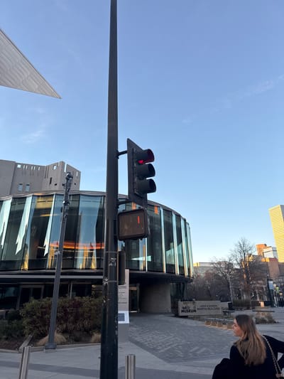 A low-angle shot of Lanny & Sharon Martin Building at the Denver Art Museum on a sunny day. A traffic light and pedestrian signal are in the foreground, with the museum's curved glass façade visible. A triangular building is seen in the background. A woman in a black coat is visible on the right side of the frame.