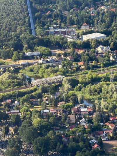 This aerial photograph captures a densely wooded hillside dotted with residential buildings and infrastructure in Ziąbki, Poland. A winding road with several vehicles ascends the steep, forested slope on the left side of the frame. Below the road, a cluster of buildings, including a long, white structure with visible signage, appears to be an industrial or commercial complex.
Further down the hillside, a body of water, likely a river, meanders through the landscape. Spanning the water is a white truss bridge, a prominent feature of the scene, with train tracks running alongside its approach. Another bridge, supporting a road, is visible further upstream.
The lower portion of the image reveals a residential area with numerous houses, many featuring red-tiled roofs, nestled among the trees. A cemetery, with rows of headstones, is visible on the far left, partially obscured by dense foliage. Overhead power lines and a transmission tower are also present, indicating the presence of electricity infrastructure. The overall scene depicts a partially developed landscape where nature and human settlement coexist.