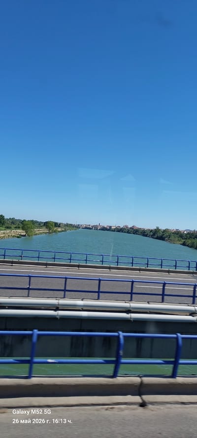 A horizontal view from a bridge in Amposta, Spain. The sky is clear blue, taking up more than half the image. The river Ebro flows alongside the bridge, with trees lining its banks. Buildings are visible in the distance. The bridge has a blue railing and gray support structures. The surface of the bridge is gray asphalt with a white stripe. The image appears to have been taken on May 26, 2026.