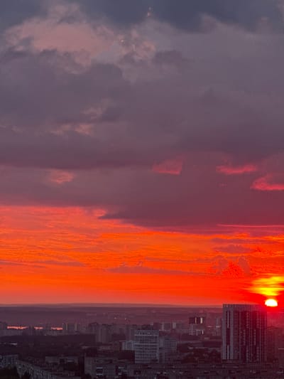 The image captures a dramatic sunset over the city of Savenki, Russia. The sky is a vibrant canvas of fiery orange and deep purple, with clouds tinged by the setting sun's intense hues. Below the spectacle of the sky, the urban landscape unfolds. Buildings of varying heights, predominantly residential blocks, stretch across the horizon. A large, modern building with a distinctive vertical pattern stands out among the others, its façade reflecting the last rays of the sun. The lower parts of the city are cast in shadow, creating a stark contrast between the illuminated sky and the darkening city. The scene is devoid of any visible people or specific actions, presenting a serene, yet powerful, depiction of an urban environment at dusk.