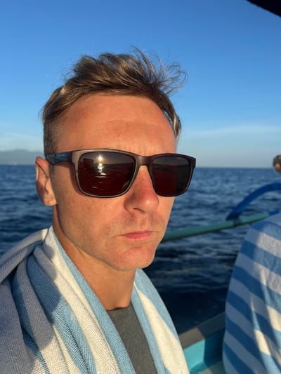 A fair-skinned man wearing sunglasses and draped with a blue and white striped towel is in a boat on the water. The sky is visible in the background. The location is specified as Seririt, Indonesia, though there is no evidence in the image to support this.