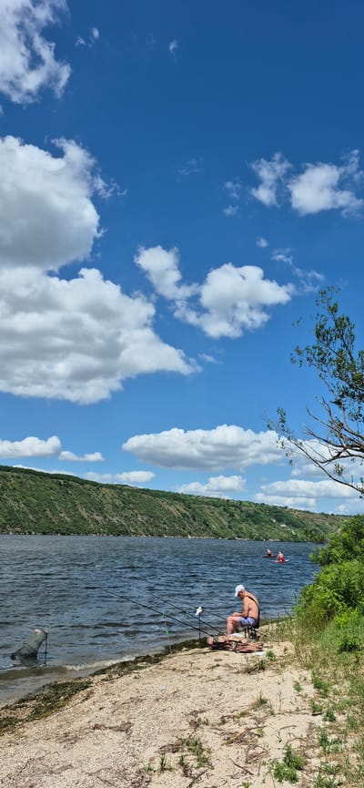 The image depicts a serene waterside scene, likely on the Nistru River near Molovata Nouă, Moldova. The sky is a vibrant blue, dotted with fluffy white clouds. In the foreground, a sandy and pebbly shore meets the dark, rippling water. A person wearing a white cap and swimwear is seated on the shore, appearing to be fishing, with several fishing rods set up beside them. A fishing net is submerged in the water to the left. In the background, a lush, green, tree-covered hillside rises from the opposite bank of the river. Further out on the water, two individuals are visible on paddleboards. The overall atmosphere is one of peaceful outdoor recreation.
