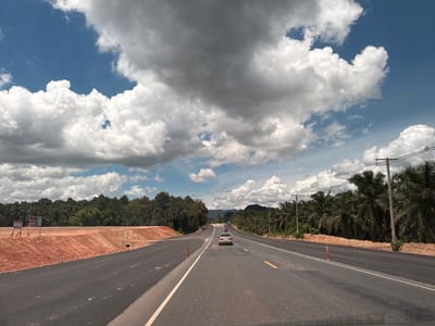 The image depicts a view from a moving vehicle on a multi-lane highway under construction, possibly in the vicinity of Plai Phraya, Thailand. The road is paved, with newly laid asphalt visible. Orange construction cones are placed along the lanes, and on the left side, an area of freshly dug, reddish-brown soil indicates ongoing earthworks. Signs are posted on the left side of the road, near the construction area. A silver sedan is driving on the highway ahead of the viewer. The highway is flanked by lush green tropical vegetation, primarily palm trees, and dense foliage. In the background, rolling hills and mountains are visible under a dramatic sky filled with a mix of white and dark, heavy clouds. Power lines supported by wooden poles run alongside the right side of the road. The lighting suggests it is daytime, with some areas of the sky brightly lit by the sun and others obscured by clouds.