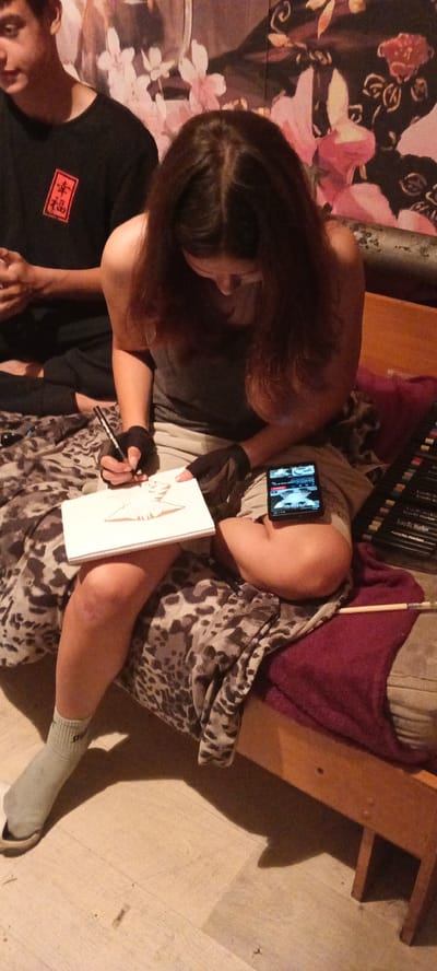The image depicts a young person sitting on a bed and drawing in a sketchbook. The person has long, dark hair and is wearing a dark gray tank top and light-colored shorts. They are using a black pen to draw on a white sketchbook that is resting on their lap. A cell phone is propped up against their leg, displaying a video. To the right of the person, a set of colored markers is neatly arranged in a box. The bed is covered with a patterned blanket featuring a leopard print and a burgundy throw. In the background, a wallpaper with a floral design is visible. To the left, another person, a young male with short dark hair, is partially visible, wearing a black t-shirt with a red rectangular emblem on it. The floor appears to be made of light-colored wood. The scene is illuminated by artificial lighting, casting shadows on the floor. The image was captured in a residential indoor setting, likely a bedroom. The image does not contain any information that would allow for its location to be determined within Chernyavskii, Russia, or any other specific geographic location.