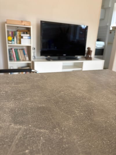 The image depicts an interior residential space, likely a living room, in Germering, Germany. The foreground features a close-up view of a textured, matte grey-brown surface, possibly a tabletop or countertop, which occupies the lower half of the frame and is in focus.
The mid-ground and background are blurred. Centrally positioned is a large, black flat-screen television with its screen off, resting on a white, low-profile media console. A small, dark object, resembling a remote control, is visible on the console to the left of the TV. To the right of the television on the console, a dark brown, abstract statuette or sculpture of a humanoid figure is present.
To the left of the television setup, a white, vertical bookshelf contains multiple rows of books, a yellow spherical object, and other indistinguishable items. A light-colored wooden box rests horizontally on top of the bookshelf.
To the right of the media console, an architectural opening reveals a glimpse into an adjacent room, possibly a kitchen, where a white appliance or counter is partially visible. The walls throughout the visible area are painted a uniform, light, neutral color, consistent with beige or off-white. No individuals are present, and no active interactions or specific actions are depicted within the scene. The lighting suggests an ambient, indoor environment.