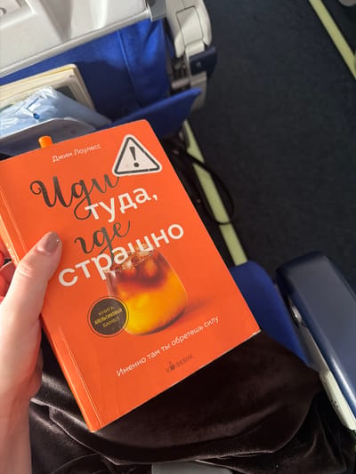 A person's hand, with lightly painted fingernails, holds an open book with an orange cover. The book displays a stylized title in Russian, which translates to "Go where it's scary." Below the title, an image of a glass filled with a dark liquid and ice is visible. A circular emblem on the book contains Russian text. The book is placed on a dark, velvety surface, possibly clothing. In the background, blue and white airplane seats are partially visible, suggesting the image was taken on an airplane. The overall lighting is soft. The location is not in the city of Guangyang District, China.