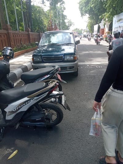 A daytime street scene in Nganjuk, Indonesia, captures a street with vehicle traffic and a pedestrian. In the immediate foreground, to the right, a person is partially visible from the mid-torso down, wearing a black long-sleeved shirt, light-colored trousers, and dark footwear, holding a transparent plastic bag containing unspecified items. To the left, two scooters are parked parallel to a striped grey and white curb; the closest is a white Honda Vario model. Adjacent to the scooters and curb, a dark green multi-purpose vehicle (MPV) is parked facing the camera. Along the left side of the street, a decorative brick wall borders a sidewalk and an area with tall trees. In the background, an asphalt road shows moderate traffic consisting primarily of motorcycles and scooters moving away from the viewer. Distant white arch-like structures with banners are visible on the right side of the road. Faint road markings reading "TAS UN" are present on the asphalt. The scene is illuminated by natural daylight.