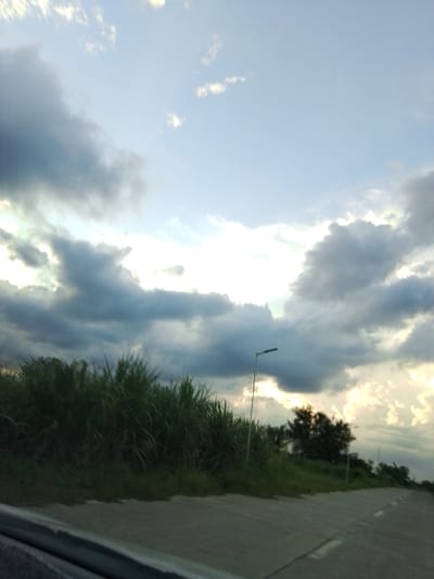 The image depicts a roadside view in Santa Cruz, Philippines, characterized by a cloudy sky and lush greenery. The foreground features a concrete road with white lane markings, suggesting a highway or major thoroughfare. To the left, a dense line of tall, green grass or reeds borders the road. A single streetlamp stands on the right side of the road, partially obscured by the vegetation. In the background, silhouetted trees are visible against a bright, hazy sky filled with dramatic clouds, some of which are illuminated by the sun breaking through. The overall impression is that of a natural landscape along a developed route, likely captured during a transitional time of day, such as late afternoon or early evening, given the quality of light. There are no people or vehicles visible in the scene.