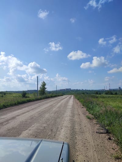 The image depicts a clear, sunny day with a bright blue sky dotted with fluffy white clouds. A dirt road stretches into the distance, flanked by green fields and trees. In the far distance, a city skyline with buildings and industrial structures, including smokestacks, is visible. The foreground shows the hood of a vehicle, reflecting the sky and surrounding environment. The scene suggests a rural or semi-rural setting outside of Staraya Uralka, Russia, with signs of both natural landscape and industrial development. There are no people or animals visible in the image.