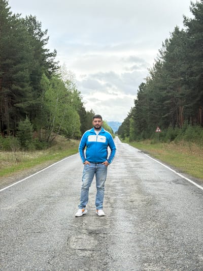 A man is standing on a road in Сизая, Russia. The man is wearing a blue Adidas jacket, jeans, and white sneakers with orange accents. The road is asphalt and has a white line marking the edge. There are trees on both sides of the road. There is a road sign with a cross on it in the distance. The sky is cloudy.