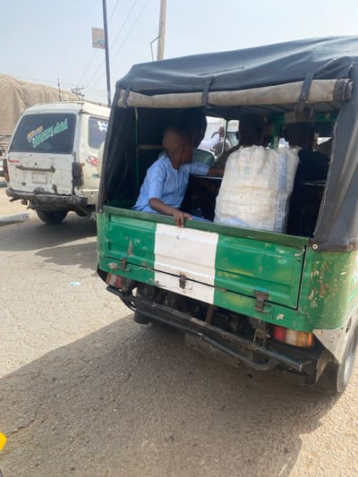 An image captured in Gobirawa, Nigeria, depicts a daytime street scene featuring a green and white three-wheeled commercial vehicle, commonly known as a keke napep, positioned prominently in the foreground. The vehicle has a black canvas roof and a rear cargo/passenger compartment painted with vertical green and white stripes. Inside, a young male child, approximately 5-7 years old, with a shaved head and wearing a light blue striped tunic, is seated on the left, looking to his right, with his hands resting on the side rail. A large white sack, bearing partial text "GOLD STAR" and "US," is centrally placed in the compartment. At least three other adult individuals are partially visible further inside the vehicle. Behind the three-wheeler, a heavily worn white van or minibus is parked or moving slowly, displaying a sticker that reads "Yummy dad" on its rear window. The vehicles are situated on an asphalt road under clear daylight conditions, casting discernible shadows. Power lines and a distant billboard are visible in the upper background.