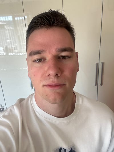 A man is shown in a close-up, slightly angled portrait. He has short, dark hair, light skin, and brown eyes. He's wearing a white t-shirt. The background consists of a white cabinet or wall with silver handles. There's no verifiable evidence that the image was taken in Bukovlak, Bulgaria.