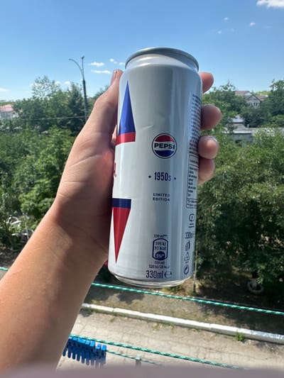 A person's hand holds a 330ml Pepsi can with a "1950s LIMITED EDITION" design against a backdrop of trees and buildings under a blue sky. The can features the Pepsi logo and nutritional information, including "395 kJ 93 kcal" and "120 kJ/28 kcal" per 100ml. The environment suggests a residential area with a street light visible in the distance. The image is taken outdoors on a sunny day.