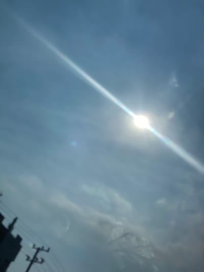 The image depicts a sky with clouds and the sun. A bright, lens-flared sun is visible in the upper right quadrant, emitting a strong diagonal ray of light across the frame. In the lower left quadrant, a dark, out-of-focus silhouette of a building and utility poles are present. The overall impression is of a slightly hazy or overcast sky, with the sun partially obscured. The location information provided, Sungai Lakum, Indonesia, suggests an outdoor urban or semi-urban environment. The image captures a moment in time, likely during daylight hours. There are no people or discernible actions visible in the scene.