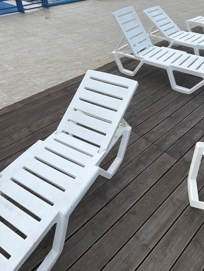 The image shows an outdoor scene with several white plastic chaise lounges arranged on a dark wooden deck. The deck is laid out with planks running diagonally from the bottom left to the top right of the frame. In the background, a tiled surface meets the deck, and beyond that, a portion of a blue structure and a blue pool edge are visible. The lighting suggests an overcast day. No people are present in the image. The scene appears to be a poolside or recreational area. The location is Yuzhnyye Koryaki, Russia.