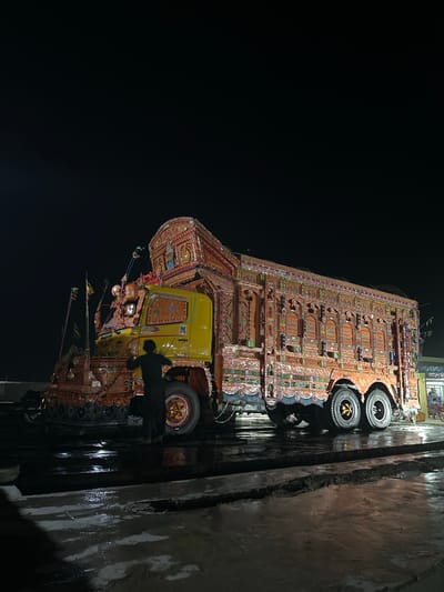 A Pakistani truck, adorned with vibrant, intricate decorations and lights, is parked at night on a wet, paved surface. A person, wearing dark clothing, stands beside the truck's front wheel, appearing to be interacting with the vehicle. The truck is heavily ornamented with colorful patterns, motifs, and religious calligraphy. The background is dark, suggesting it is nighttime, with some distant lights and structures visible. The wet ground reflects the truck's lights, creating a glossy sheen on the surface.