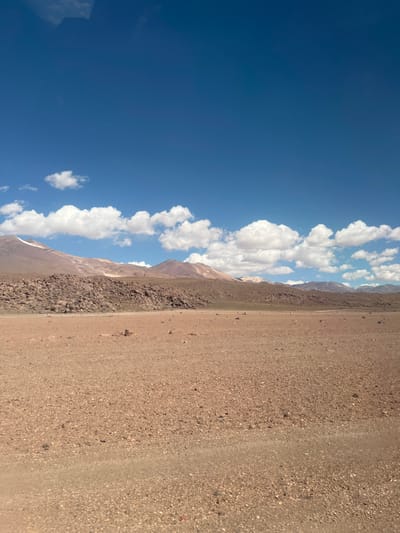 A landscape view features a clear blue sky with scattered white clouds on the left. Adjacent to the clouds are rocky mountains that transition into a vast, dry, arid terrain on the right. The ground is covered in small rocks and sand, creating a textural contrast to the smooth sky. The scene suggests a remote, desolate location under bright sunlight.