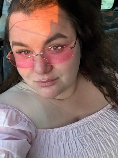 The media file shows a close-up, high-angle shot of a young person with long, dark hair, wearing pink, rimless sunglasses and a ruched pink top. The person's face is angled towards the camera, with their left eye and cheek prominently visible, illuminated by warm, orange light from what appears to be the sun. Their lips are slightly parted, and they have dark eyebrows and eyelashes. The background is blurred, showing hints of a car interior with textured upholstery and a window. The lighting suggests it might be late afternoon or early morning, with the sun casting a warm glow on the person's skin. The setting is likely inside a vehicle. There is no visible text. The overall mood is casual and personal, possibly a selfie taken while traveling.