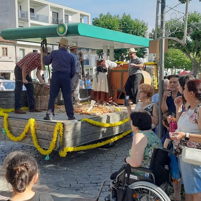The image depicts a street scene in Vila Verde, Portugal, under a bright, sunny sky. In the background, a white building with multiple balconies and a green BP gas station canopy are visible. The foreground features a wooden cart laden with corn cobs, decorated with yellow garlands. Several people are present, some dressed in traditional attire, suggesting a festival or cultural event.
In the center of the image, a group of individuals are gathered around the cart. A man in a blue plaid shirt and straw hat is bent over, working with a large basket and a tool. Another man in a similar hat and blue plaid shirt stands nearby, holding a similar tool and conversing with a woman in traditional dress. The woman wears a white blouse with a brown vest and a long, patterned skirt. To the right, a man in a checkered shirt and straw hat is speaking to someone.
Further to the right, a woman in a wheelchair is visible, facing away from the camera and holding a smartphone. Another woman, wearing a floral-patterned top and denim shorts, stands next to her, holding a white purse. Various other onlookers are scattered throughout the scene, some interacting and others observing. The ground is paved with cobblestones. The overall atmosphere appears to be one of a lively community gathering.