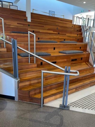 This image captures a modern, indoor architectural space, likely a university building or a public facility in Kelvin Grove, Australia. The central feature is a wide, amphitheater-style staircase or terraced seating area, constructed from warm-toned, polished wooden planks arranged in varying widths.
The wooden steps are designed to accommodate seating, with several elongated, oval-shaped cushions or backrests integrated directly into the steps. These padded sections are dark grey with a subtle patterned texture. Small, black rectangular inserts, possibly electrical outlets or data ports, are embedded intermittently into the wooden surfaces.
To the right, a contemporary handrail system with shiny silver metal (likely stainless steel) tubing curves upwards, providing support along the steps. This railing has a distinctive U-shaped base where it meets a darker, speckled floor area. Further up and to the right, a simpler grey metal railing is bolted into a lighter-colored wall or floor section, distinct from the main handrail. On the far left, a light-colored wall ascends alongside the wooden structure, meeting a ceiling area with ample natural light streaming in from windows, suggesting an upper floor or atrium. Another set of silver metal handrails, integrated with glass panels, is visible further back, indicating a more conventional staircase.
The space is currently devoid of people, suggesting a quiet moment or an off-peak time. The bright natural light indicates it is daytime. Visible text includes a green and white "EXIT" sign high on the left wall. Below it, a yellow and black striped tape barrier is present, possibly indicating a temporary closure or warning. A faint white rectangular sign, partially obscured, appears to display "WASH YOUR HANDS," suggesting a focus on public health. The overall impression is one of a clean, functional, and aesthetically pleasing public gathering or transit area.