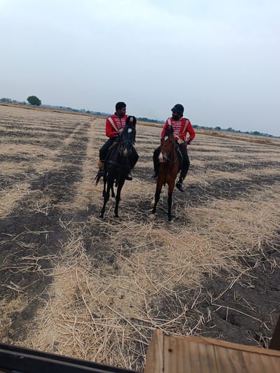 In an open field in Kottalapalli, India, two adult males are mounted on horseback under an overcast sky. The ground consists of dark soil interspersed with dry, light-brown crop stubble and visible furrows. The individual on the left rides a black horse with a white blaze and wears a red military-style jacket with white horizontal stripes, black pants, and a dark helmet. He faces partially towards the second individual. The individual on the right rides a brown horse with a white blaze, white muzzle, and white socks on its front hooves. He wears a similar red military-style jacket, black pants, and a dark helmet. Both individuals appear to be conversing. In the distant background, a flat horizon features scattered trees and faint vegetation. A dark, wooden structure is partially visible in the bottom foreground, suggesting the vantage point of the photographer.